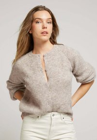 Cardigan en tricot en fibres beige douces, avec un col rond, une fermeture à boutons et des manches courtes. Texturé avec des détails en ribbing vertical.