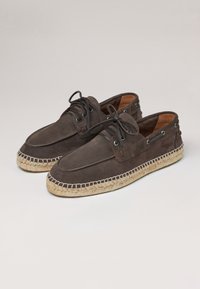 Zapatos de barco de ante marrón con una suela de esparto texturizada, puntera redondeada y cordones decorativos, que cuentan con ojales de metal y acentos de costura.