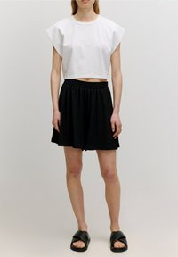 T-shirt bianca corta con maniche corte a tesa; abbinata a una gonna nera plissettata e sandali neri slip-on. Tessuto strutturato con cintura elastica.