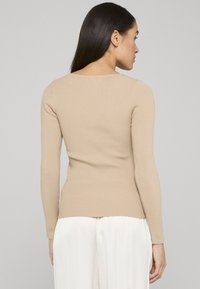 Femme aux longs cheveux noirs portant un haut beige côtelé à manches longues ajusté et un pantalon blanc ample, debout de dos sur un fond uni.