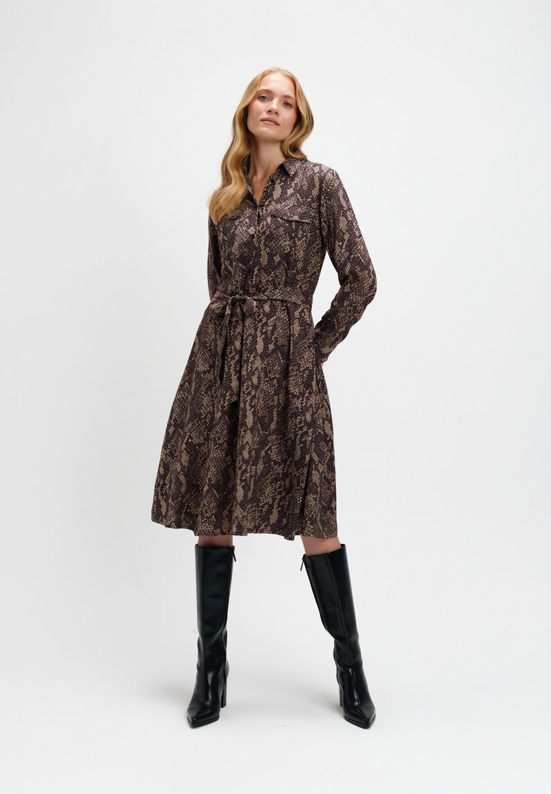 Sukienka z długim rękawem w brązowym i beżowym wzorze węża, z kołnierzykiem, paskiem w talii oraz dwoma kieszeniami na piersiach. Dopasowana z czarnymi botkami za kolano.