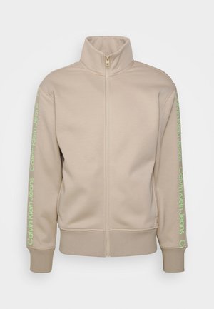 Veste beige zippée avec col montant, manches et ourlet côtelés, arborant un texte vert sur les manches avec "Calvin Klein Jeans". Texture de tissu douce.