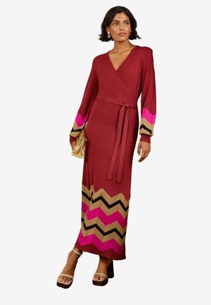 Kvinde iført en lang rød slå-om-kjole med pink, beige og sorte chevronmønstre, der holder en gylden taske og har beige plateau-sandaler på.