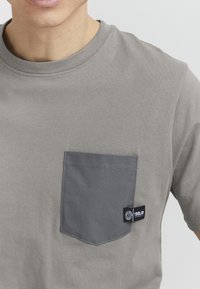 Gray cotton t-shirt with a round neckline and a contrasting gray patch pocket on the left chest. Features a small brand tag on the pocket.