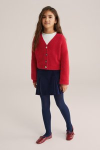 Raudona puri megztinė su V formos iškirpte ir juodais gėlių mygtukais, dėvima virš balto marškinėlio, kartu su tamsiai mėlyna sijonu ir tamsiai mėlynomis pėdkelnėmis, užbaigta raudonais batais.