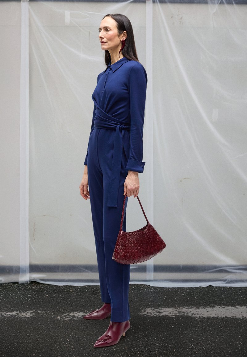 Marineblå slå-om-jumpsuit med lange ærmer og et bindebælte, kombineret med en bordeaux fletpose og matchende bordeaux spidse sko.