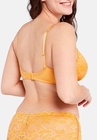 Sujetador de encaje amarillo con tirantes ajustables y cierre de gancho y ojo. Patrón floral decorativo con una textura suave y lisa.