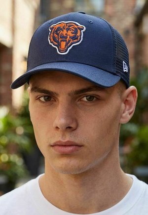 Junger Mann mit heller Haut und kurzen Haaren, der eine marineblaue Mütze mit einem orangefarbenen Bärenlogo trägt, im Freien vor unscharfem grünen Hintergrund.