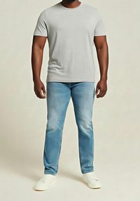 Mann trägt ein schlichteres hellgraues T-Shirt mit kurzen Ärmeln, hellblaue Jeans und weiße Turnschuhe, steht vor einem neutralen Hintergrund.