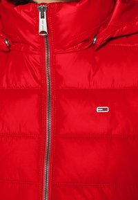 Czerwona kurtka puchowa z zamkiem błyskawicznym z przodu, pikowanym wzorem i naszywką logo. Materiał wydaje się błyszczący, ma gładką teksturę i izolacyjną konstrukcję.