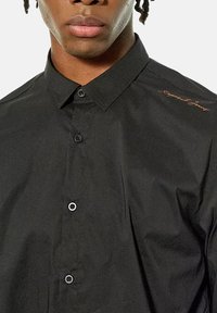 Zwarte blouse met puntkraag, voorzien van een subtiele geborduurde logo in koper op de linkerschouder en een gladde textuur.
