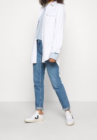 Weißes Hemd über einem hellblauen Pullover, kombiniert mit hellblauen Jeans und weißen Sneakers mit schwarzen und roten Akzenten.