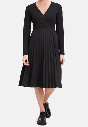 Kvinde iført en sort kjole med lange ærmer og en plisseret nederdel samt sorte blanke platformsko, stående mod en ensfarvet baggrund.