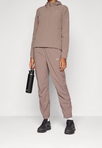 Chaqueta deportiva color marrón claro con capucha y detalles reflectantes, combinada con pantalones a juego y zapatillas negras, sosteniendo una botella de agua negra.