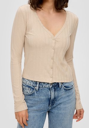 Kvinde iført en beige ribstrikket knaplukket langærmet top kombineret med højtaljede blå denimjeans, afkortet ved taljen.