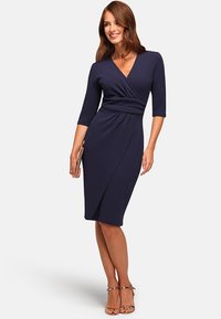 Femme souriante, portant une robe enveloppante bleu marine jusqu'au genou avec des manches trois-quarts, tenant une pochette beige et portant des talons dorés à lanières.
