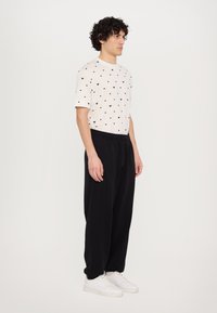White t-shirt with small black heart pattern, paired with black sweatpants, and white sneakers. Model shown in profile view.