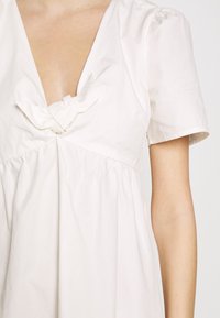 Robe blanche à manches courtes avec un décolleté en V profond et un détail de nœud en tissu sur la poitrine, montrant le haut du torse et le bras droit du porteur.