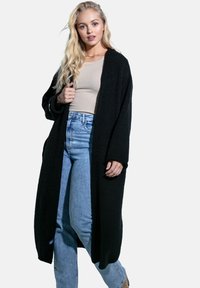 Długi czarny kardigan wykonany z teksturowanej dzianiny. Posiada luźne, szerokie rękawy oraz boczne kieszenie, noszony na beżowej bluzce i niebieskich dżinsach.
