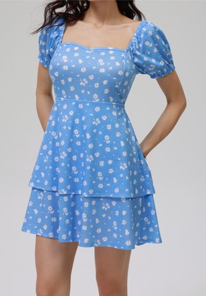 Kort blå kjole med hvidt tusindfrydmønster, puffede korte ærmer, firkantet udskæring og udvidet lagdelt nederdel.