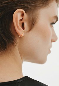 Pendiente de aro dorado en forma de media luna llevado en la oreja izquierda, con cabello corto castaño y textura de piel suave, todo contra un fondo neutro.