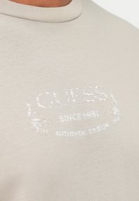 Béžové tričko s okrúhlym výstrihom a bielym embosovaným textom "GUESS," "Since 1981," a "Authentic Design" na hrudi.