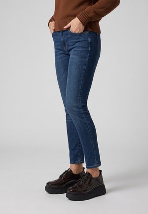 Person iført blå slim-fit jeans, brun sweater og mørkebrune plateau sko, stående med hænderne i lommerne mod en ensfarvet baggrund.