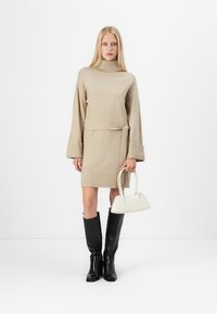 Beige stickad klänning med hög krage, bälte i midjan och vida ärmar. Tillsammans med knähöga svarta stövlar och en liten vit handväska.