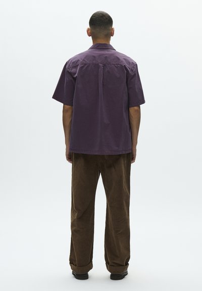 Man wearing a loose, short-sleeve purple shirt and brown pants, standing facing away on a plain light background.