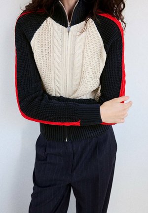 Persona que lleva un suéter de punto negro y blanco con cremallera y rayas rojas en las mangas, combinado con pantalones oscuros de rayas finas, brazos cruzados.