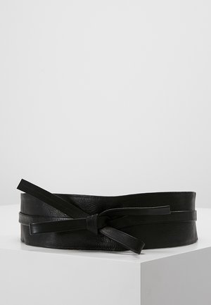 Zwarte brede leren riem met een dunne leren riem gestrikt in een knoop rond het midden, weergegeven op een witte ondergrond.