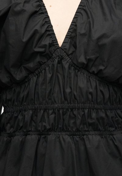 Μαύρο φόρεμα από ραμμένο ύφασμα με βαθύ V-λαιμόκοψη, ελαστικές πιέτες στο στήθος και τη μέση, που δείχνει ανοιχτό χρώμα δέρματος στο ντεκολτέ.