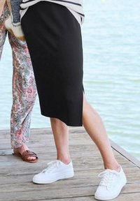 Jupe noire mi-longue avec fente latérale, associée à des baskets blanches texturées sur une plateforme en bois près de l'eau. Des motifs sur le pantalon adjacent sont visibles.