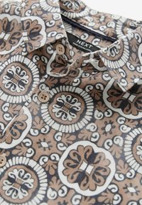 Camisa de cuello en marrón, negro y blanco con patrones florales circulares. Presenta botones de plástico y una etiqueta marcada como "NEXT". Tejido texturizado.