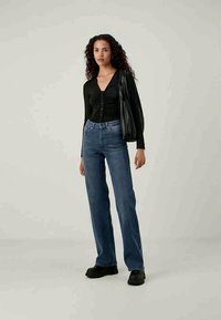 Camisa preta de mangas longas e botões com franzido, combinada com jeans flare azul de cintura alta. A modelo carrega uma bolsa preta e usa sapatos pretos.