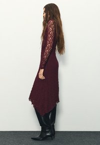 Burgunderrotes Spitzenkleid mit langen Ärmeln und einem asymmetrischen Saum, kombiniert mit kniehohen schwarzen Lederstiefeln. Ganzflächiges strukturiertes Blumendesign.