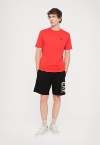 Raudona trumpomis rankovėmis T-shirt su juodu logotipu, derinama su juodomis šortais, kuriuose yra baltas prekės ženklas. Modelis dėvi šviesius sportinius batus ir kulkšninius džinsus.