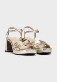 Sandales à talons métalliques dorés avec un design à brides croisées, une bride de cheville et un talon bloc. Texture lisse et finition brillante, semelle intérieure rembourrée.
