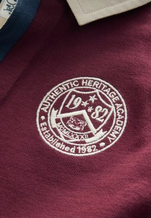 Mørkerød polo-shirt med en cremefarvet krave. Har et hvidt broderet emblem med "Authentic Heritage Academy," grundlagt i 1982.