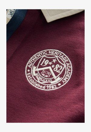 Bordowa koszulka polo z kremowym kołnierzykiem. Posiada białą haftowaną naszywkę z napisem "Authentic Heritage Academy", założoną w 1982 roku.