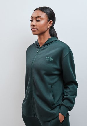 Femme aux cheveux foncés attachés, portant un sweat à capuche zippé vert foncé avec le texte "DASH AND STARS", mains dans les poches, tournée vers la gauche sur fond uni.