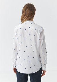Wit shirt met blauwe florale borduursels, knoopsluiting kraag, lange mouwen en een gebogen zoom. Zacht materiaal met gedetailleerde stiksels.