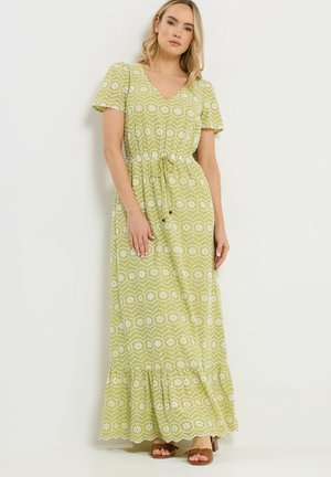 Frau in einem hellgrünen, kurzärmeligen Maxikleid mit weißen Blumenmustern und einem Kordelzug in der Taille, stehend vor einem schlichten Hintergrund.