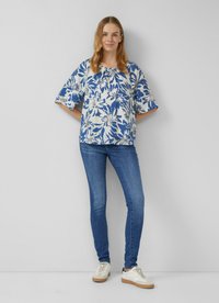 Blumenbluse in Blau und Weiß, mit kurzen Ärmeln und einem runden Ausschnitt. Kombiniert mit eng anliegenden blauen Jeans und weißen Turnschuhen.