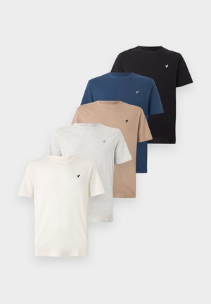 Homme portant un t-shirt noir à manches courtes, un pantalon beige et des baskets blanches, debout avec une main dans la poche devant un fond blanc uni.