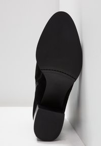 Semelle de chaussure à talon haut noire avec finition lisse, inclinée contre un mur d'angle et le sol sur un fond gris clair.