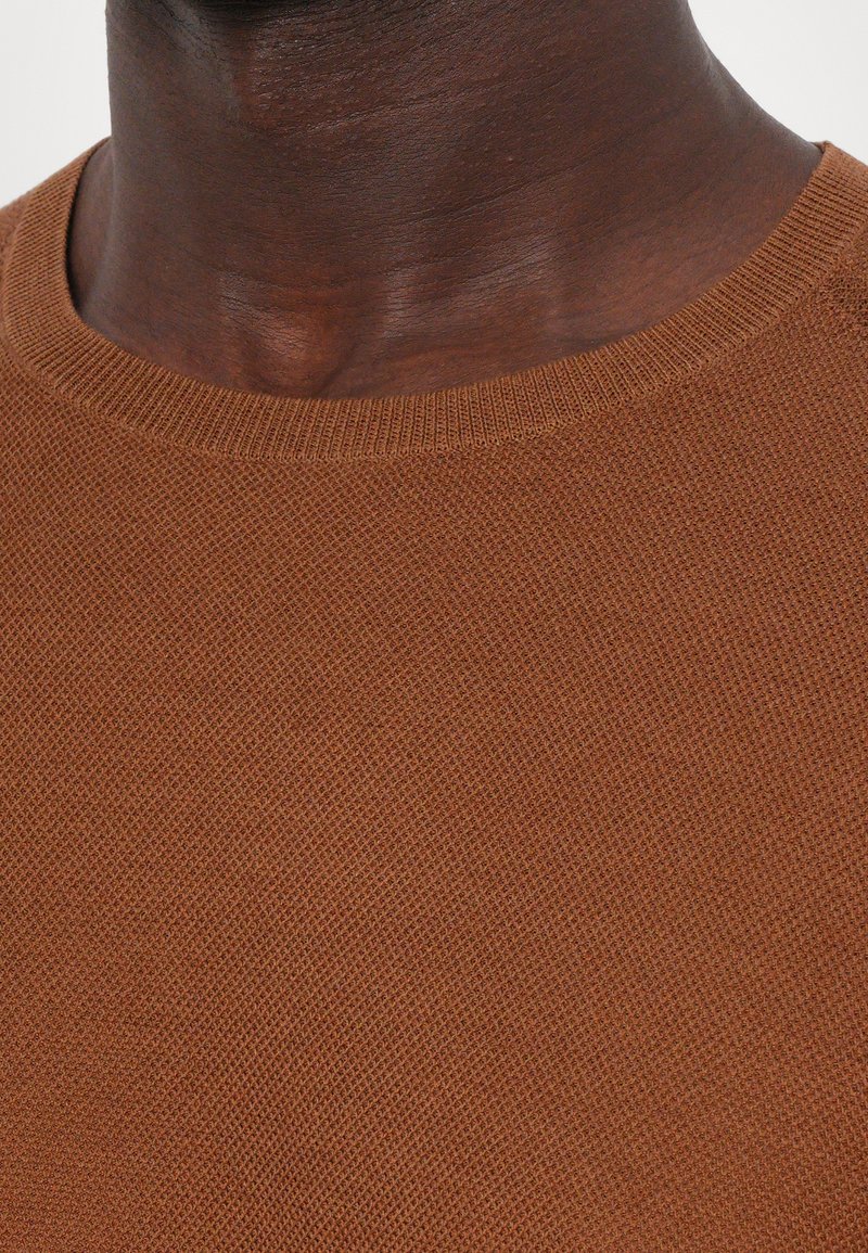 Brun stickad tröja med rund halsringning, med en strukturerad väv och subtila ribbstickade detaljer längs kragen.