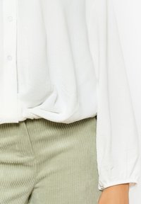 Blusa bianca con bottoni realizzata in un tessuto morbido e leggero, caratterizzata da maniche lunghe e arricciate. Abbinata a pantaloni a costine verdi oliva.