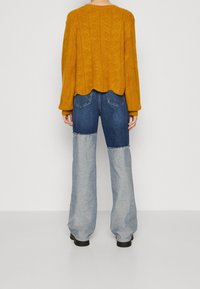 Person, der einen senfgelben, kabelgestrickten Pullover und weitgeschnittene Jeans in zwei Blautönen trägt, steht mit dem Rücken zur Kamera.