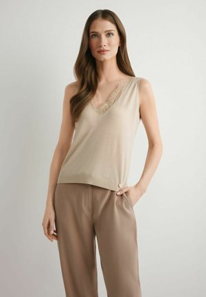 Kobieta z długimi brązowymi włosami, ubrana w beżową bezrękawnik z koronkowym wykończeniem i luźne brązowe spodnie, stojąca z jedną ręką w kieszeni.